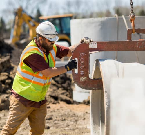 A construction worker in a safety vest and hard hat guides a large concrete pipe into place at a worksite using a metal lifting hook. Excavation equipment is visible in the background.