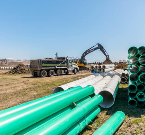 A construction site with green and gray large pipes stacked in the foreground, a dump truck and an excavator working in the background under a clear blue sky.