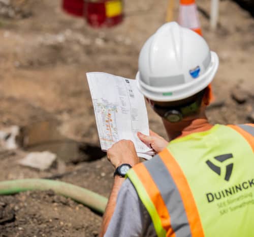 A construction worker wearing a hard hat and a Duininck safety vest reviews a site plan at a construction area with dirt, hoses, and traffic cones visible.