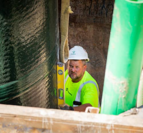 A worker in a neon yellow shirt and white hard hat uses a spirit level while working next to large green pipes and a cylindrical structure at a construction site. - December 9, 2021