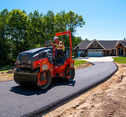 A worker operates an orange road roller to smooth freshly laid asphalt on a driveway leading to a house surrounded by greenery and trees under a clear blue sky.