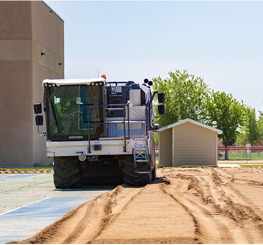 A large construction vehicle levels a dirt surface near a small shed and a building, with trees and a clear sky in the background.