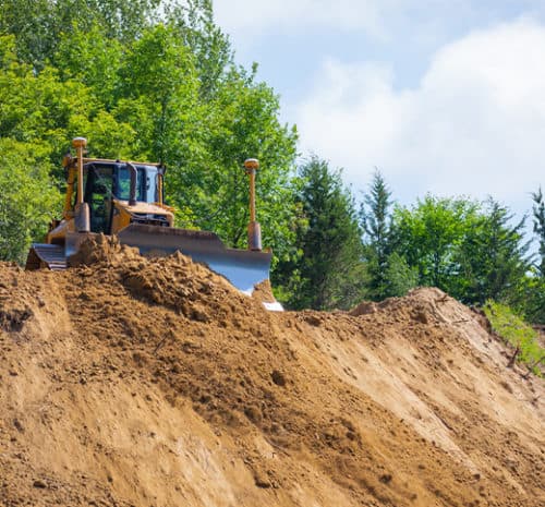 A bulldozer pushes a large mound of dirt at a busy construction site, with green trees and a partly cloudy sky in the background.