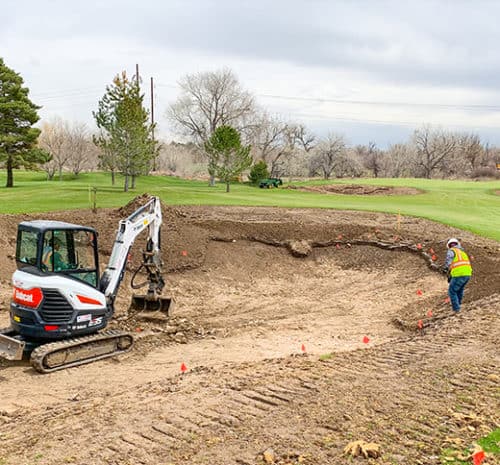 A construction worker in a safety vest stands near a partially dug pit on a golf course, while a small excavator is parked nearby. Orange flags mark the area, and trees and grass surround the site.
