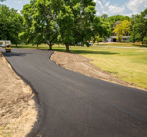 A freshly paved asphalt driveway curves through a green, grassy yard with trees and a house visible in the background under a sunny sky. A construction vehicle is parked near the start of the driveway.