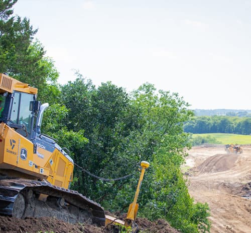 A yellow bulldozer works on a dirt slope near trees at a construction site, overlooking earthmoving equipment in the distance under a clear sky.