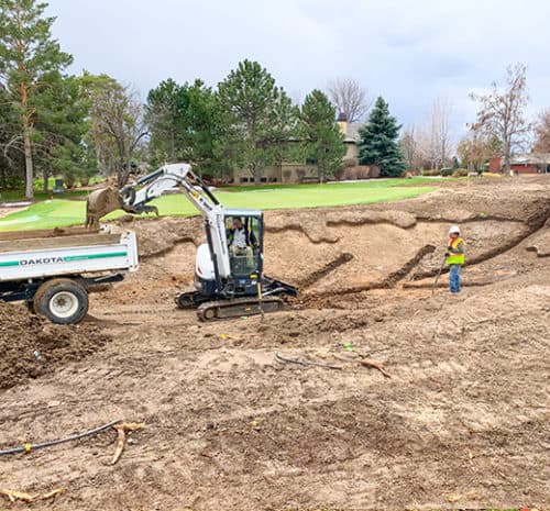 A construction worker operates a small excavator, loading dirt into a dump truck, while another worker in a yellow vest and hard hat observes on a construction site near a grassy area with trees and houses in the background.