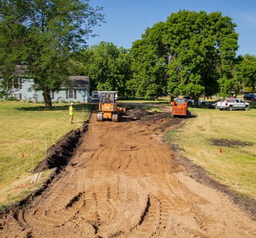 Construction workers use heavy machinery to clear and grade a dirt path on a grassy area near a house and several trees, preparing the ground for a new driveway or road on a sunny day.