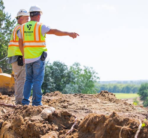 Two construction workers in safety vests and helmets stand on a dirt mound at a construction site. One points into the distance while the other looks on, with trees and open land visible behind them.