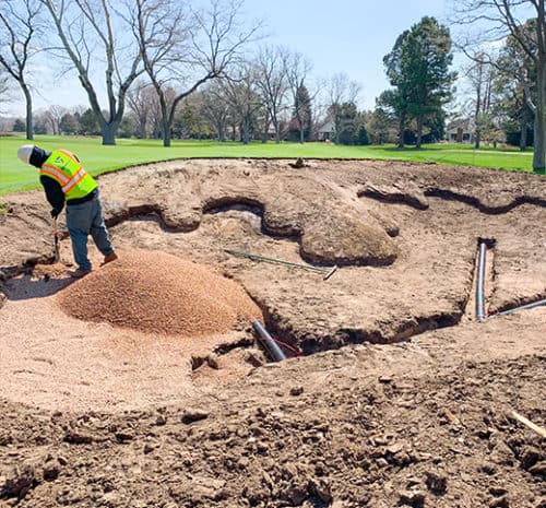 A worker in a yellow safety vest spreads gravel in a large, partially constructed sand bunker at a golf course. Piles of gravel, exposed pipes, and bare trees are visible in the background.