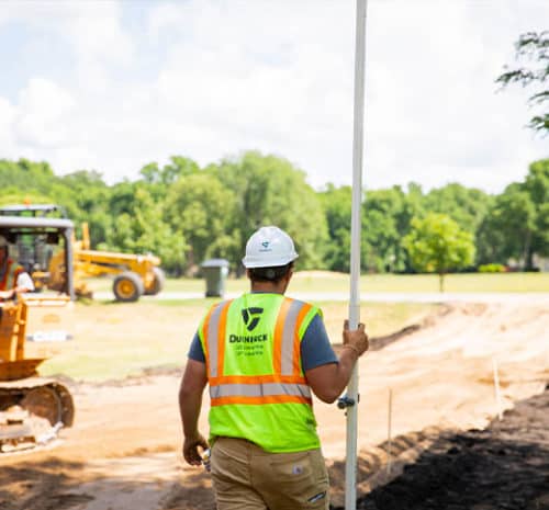 A construction worker in a safety vest and hard hat walks on a dirt construction site holding a tall pole, with construction equipment and greenery visible in the background.