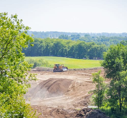 A bulldozer levels dirt on a large, open construction site, surrounded by green trees and grassy fields under a clear sky, highlighting the busy world of construction.