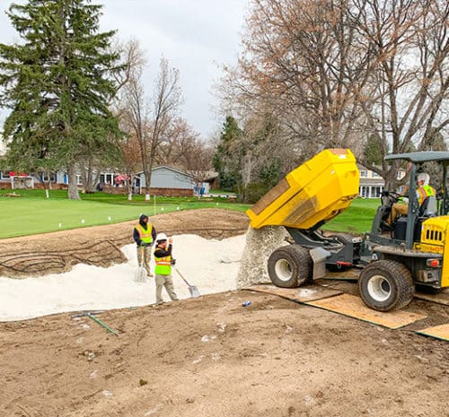 Use shovels to spread sand in a golf course bunker, while a yellow dump truck unloads more sand. Trees and houses are visible in the background.