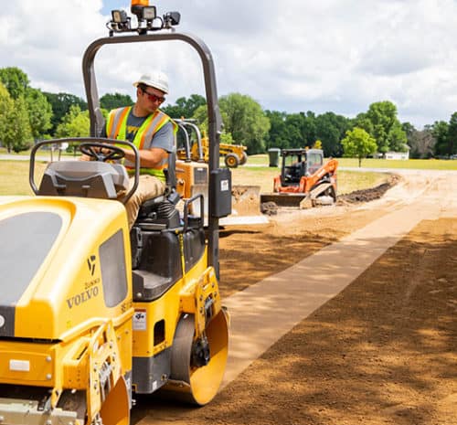 A construction worker wearing a safety vest and helmet operates a yellow Volvo road roller, compacting a dirt path at an outdoor construction site with trees and other equipment in the background.