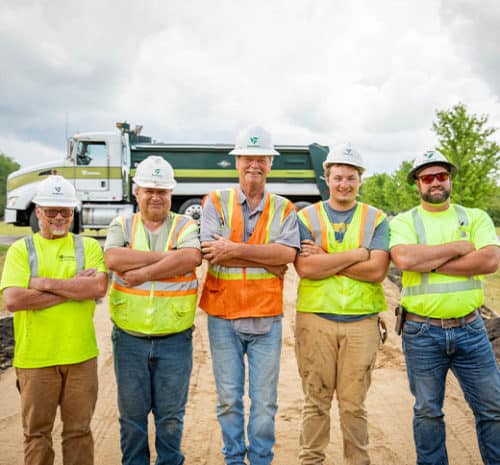 Five construction workers wearing safety vests and helmets stand side by side with arms crossed, smiling at the camera. A dump truck is visible in the background on a cloudy day.
