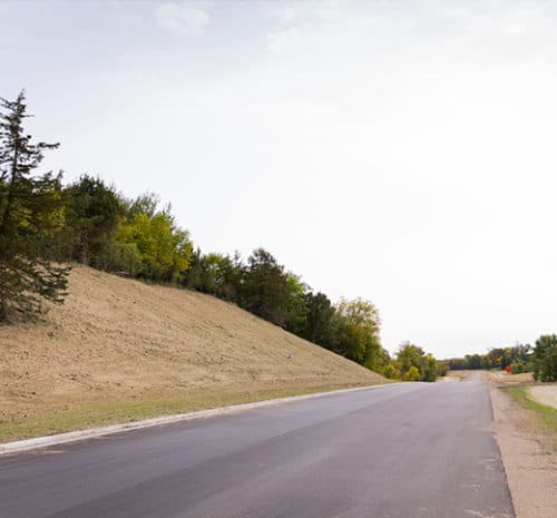 A freshly paved road, the result of recent construction, runs alongside a sloped, grassy embankment with trees on the left under a cloudy sky. The scene is quiet, with no vehicles or people visible.