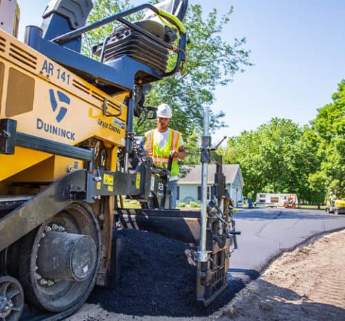 A construction worker operates an asphalt paving machine, laying new pavement on a residential street lined with trees and houses on a sunny day.