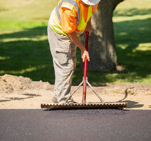 A construction worker in a reflective vest smooths fresh asphalt with a large rake on a sunny day, with grass and a tree visible in the background.