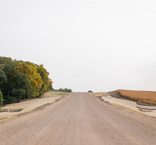 A dirt road under a pale, overcast sky stretches uphill into the distance, bordered by green and yellow trees on the left and a harvested field on the right—hinting at recent construction in the area.