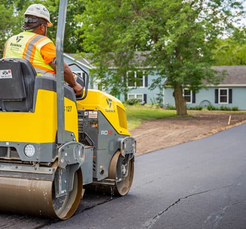 A construction worker operates a yellow asphalt roller, smoothing fresh pavement on a residential street lined with green trees and a light blue house in the background.
