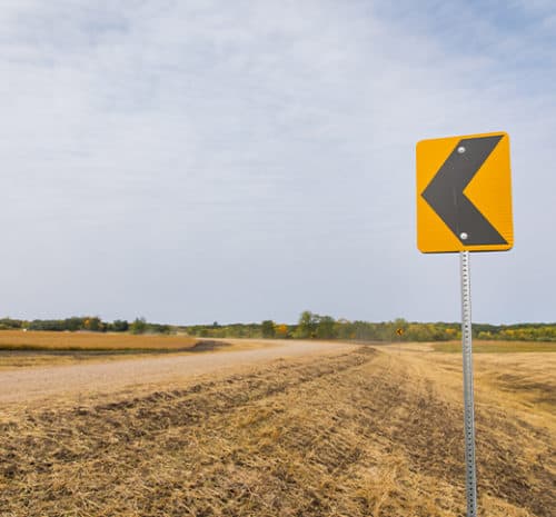 A yellow road sign with a black left arrow stands beside a rural dirt road that curves to the left, surrounded by fields and trees under a cloudy sky, hinting at possible construction ahead.