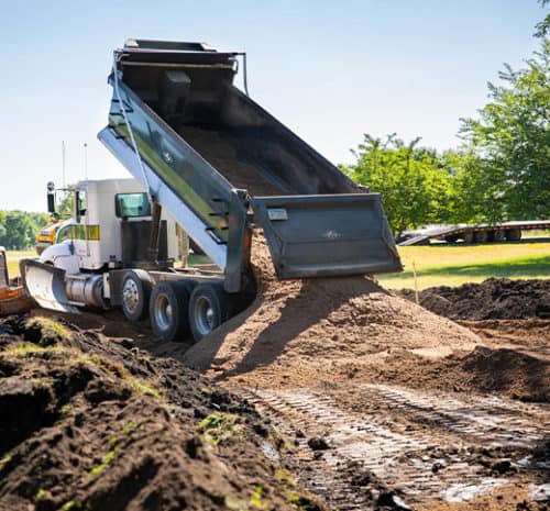 A dump truck unloads a large pile of dirt onto a construction site, with tracks visible on the ground and green trees in the background under a clear blue sky.