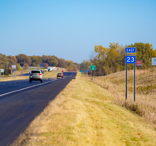 A highway with two cars driving along it, bordered by grassy shoulders and trees. A blue road sign on the right reads East Minnesota 23 under a clear, blue sky.