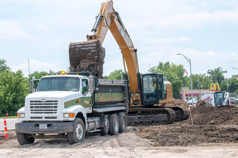 A large excavator loads dirt into a dump truck at a construction site on a sunny day, with trees, a McDonalds sign, and other construction vehicles visible in the background.