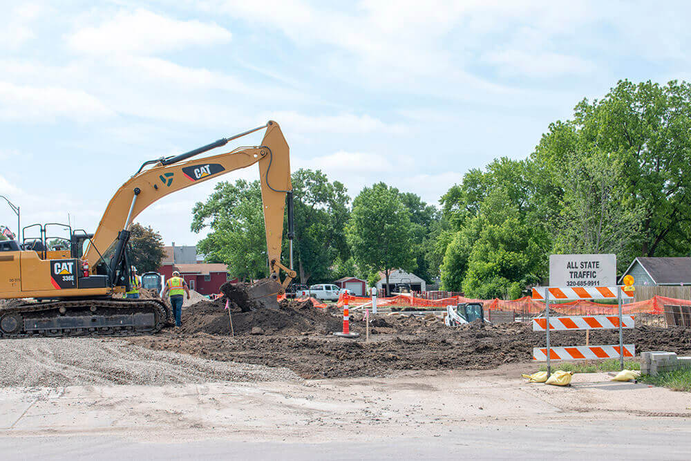 A yellow excavator moves dirt at a construction site surrounded by orange safety fencing. A sign reads All Site Traffic with an arrow. Trees and several parked vehicles are visible in the background.