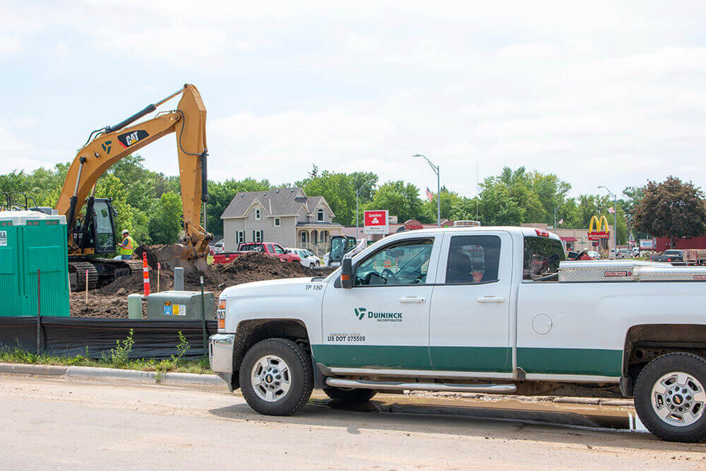 A construction site with a backhoe digging near a portable toilet. Workers in safety vests are present. A Dunnick company pickup truck is parked in the foreground. Houses and a McDonalds restaurant are visible in the background.