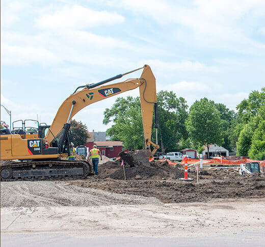 A yellow excavator is digging and moving dirt at a construction site as two workers in safety vests stand nearby. There are trees and houses in the background under a partly cloudy sky.