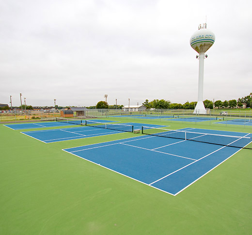 Four empty blue and green outdoor tennis courts with a tall water tower labeled Clara City in the background, under a cloudy sky. Trees and other park facilities are visible in the distance.