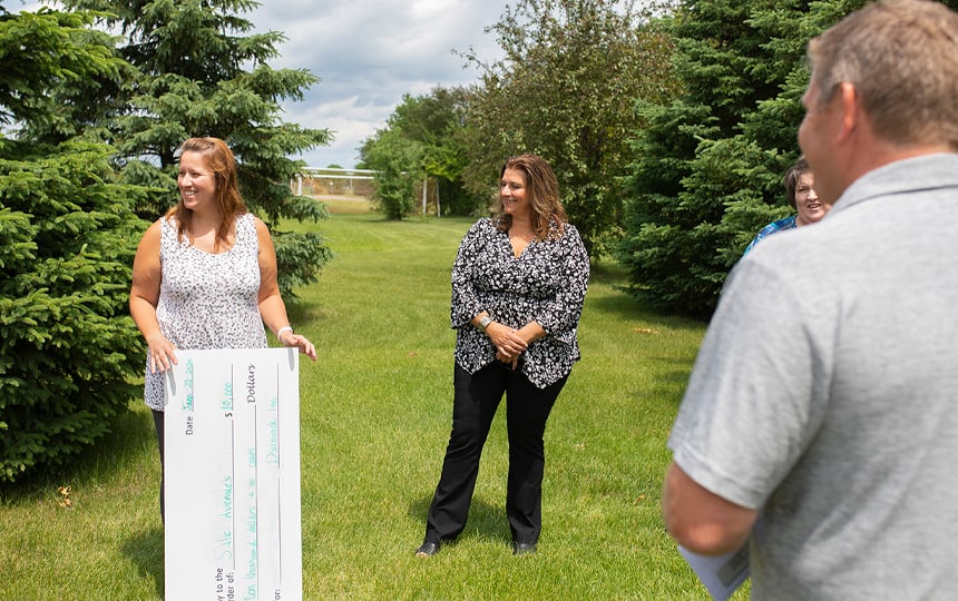 A woman holds an oversized check and smiles while standing outside on grass with three other adults, surrounded by trees and under a partly cloudy sky.