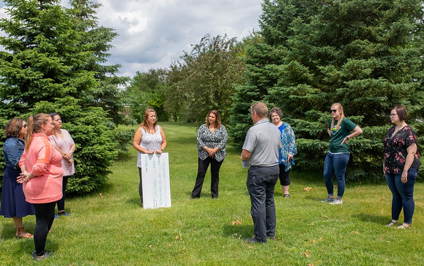 A group of nine adults stands in a loose circle on green grass between evergreen trees, outdoors on a partly cloudy day. One woman holds a large white poster board with text. The group appears to be engaged in conversation.