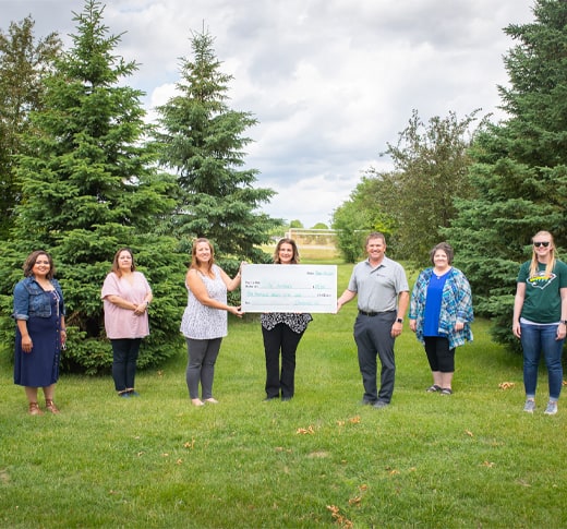 Seven people stand on grass in a park-like setting, spaced apart, with four women in the center holding a large presentation check. Pine trees and cloudy sky are in the background.