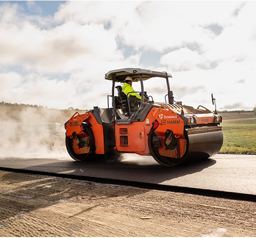 A worker operates an orange HAMM road roller, compacting hot asphalt on a rural road under a partly cloudy sky, with fields visible in the background.