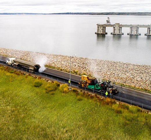 Aerial view of road construction vehicles paving a road beside a body of water, with workers in safety vests nearby and a rocky embankment separating the road from the water.