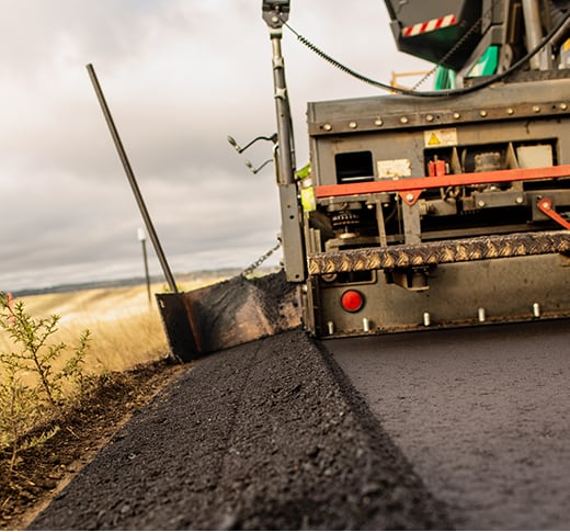 A close-up view of a road paving machine laying fresh asphalt on a rural road, with fields and cloudy sky in the background.