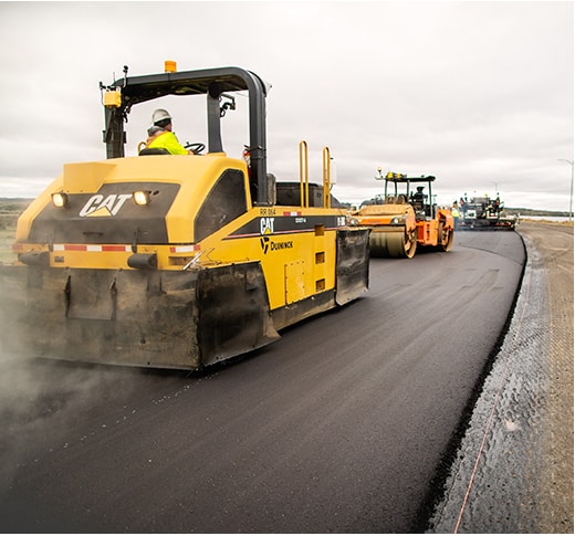 A construction worker operates a large yellow asphalt roller, smoothing freshly laid pavement on a road under cloudy skies, with more construction vehicles following behind.