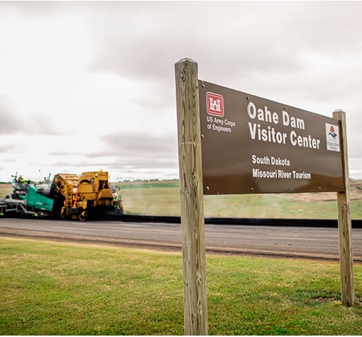 A brown sign reading Oahe Dam Visitor Center, South Dakota, Missouri River Tourism stands in front of road construction equipment on a grassy area under a cloudy sky.
