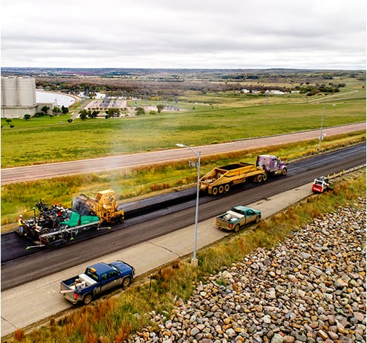 An aerial view of a road construction site with vehicles paving a road. Trucks and construction equipment are working, while cars are parked nearby. Green fields and distant buildings are visible under a cloudy sky.