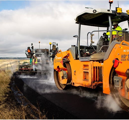 Construction workers operate heavy machinery laying and compacting fresh asphalt on a road. Steam rises from the hot surface, while workers in safety gear oversee the process under a cloudy sky.