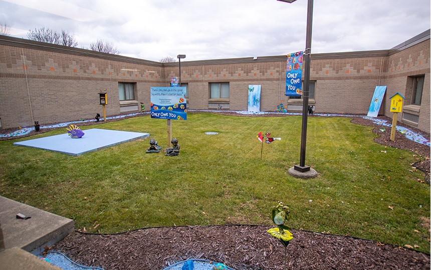 A small grassy courtyard enclosed by a brick building, decorated with signs, banners reading Only One You, painted rocks, statues, and a few birdhouses, with a cloudy sky overhead.