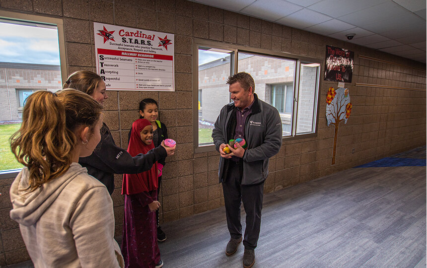 A man hands out colorful objects to three smiling children in a school hallway near a window. An adult stands beside the children. A sign on the wall reads Cardinal S.T.A.R.S. in red letters.