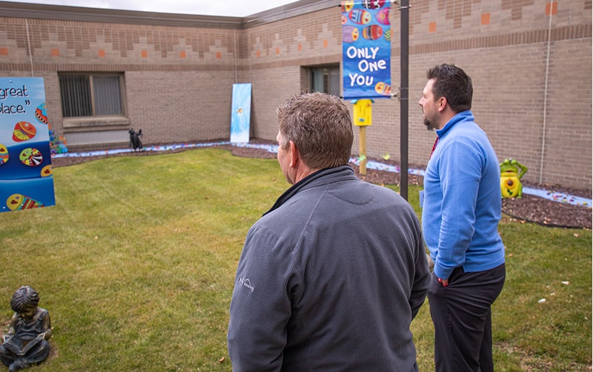 Two men stand outside on a grassy area near a building, facing away. Decorative banners, statues, and colorful decorations are placed around the yard. The scene appears calm and contemplative.