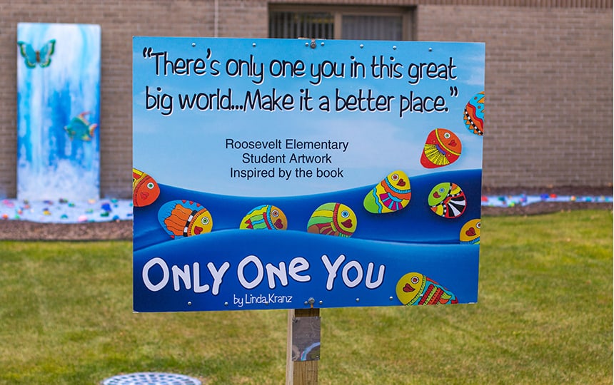 A colorful outdoor sign reads: Theres only one you in this great big world... Make it a better place. It features painted fish rocks and mentions Roosevelt Elementary student artwork inspired by the book Only One You.