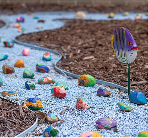 A garden path made of blue gravel winds through mulch, decorated with colorful painted rocks and a painted fish sculpture on a stake.