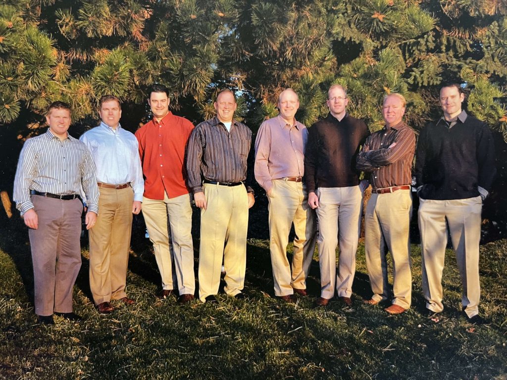 Group of eight men posing in front of trees outdoors